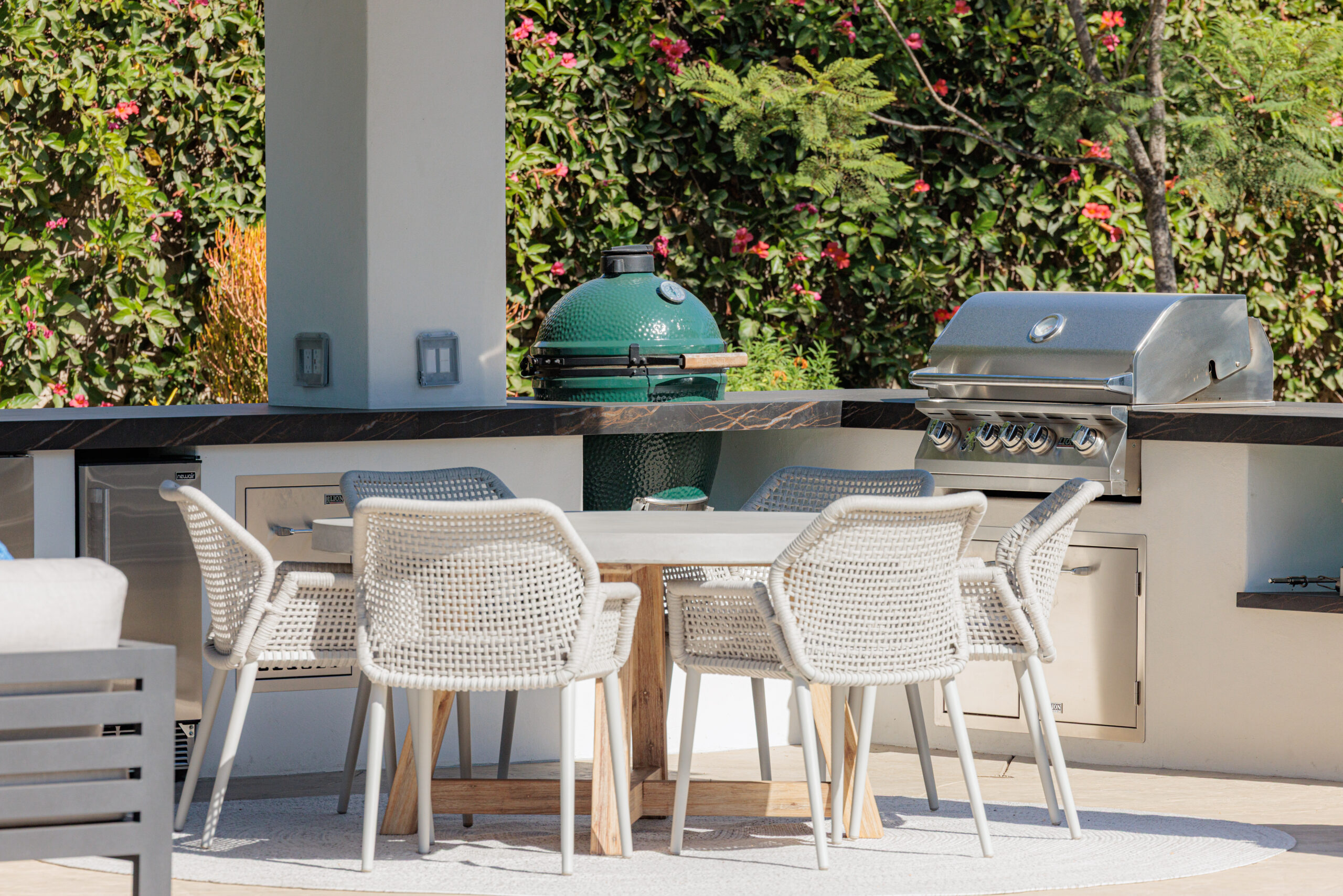 Outdoor kitchen with round modern dining table and chairs, and custom built masonry kitchen island with white stucco, 32" stainless steel grill, green egg, and landscape hedge in the background.