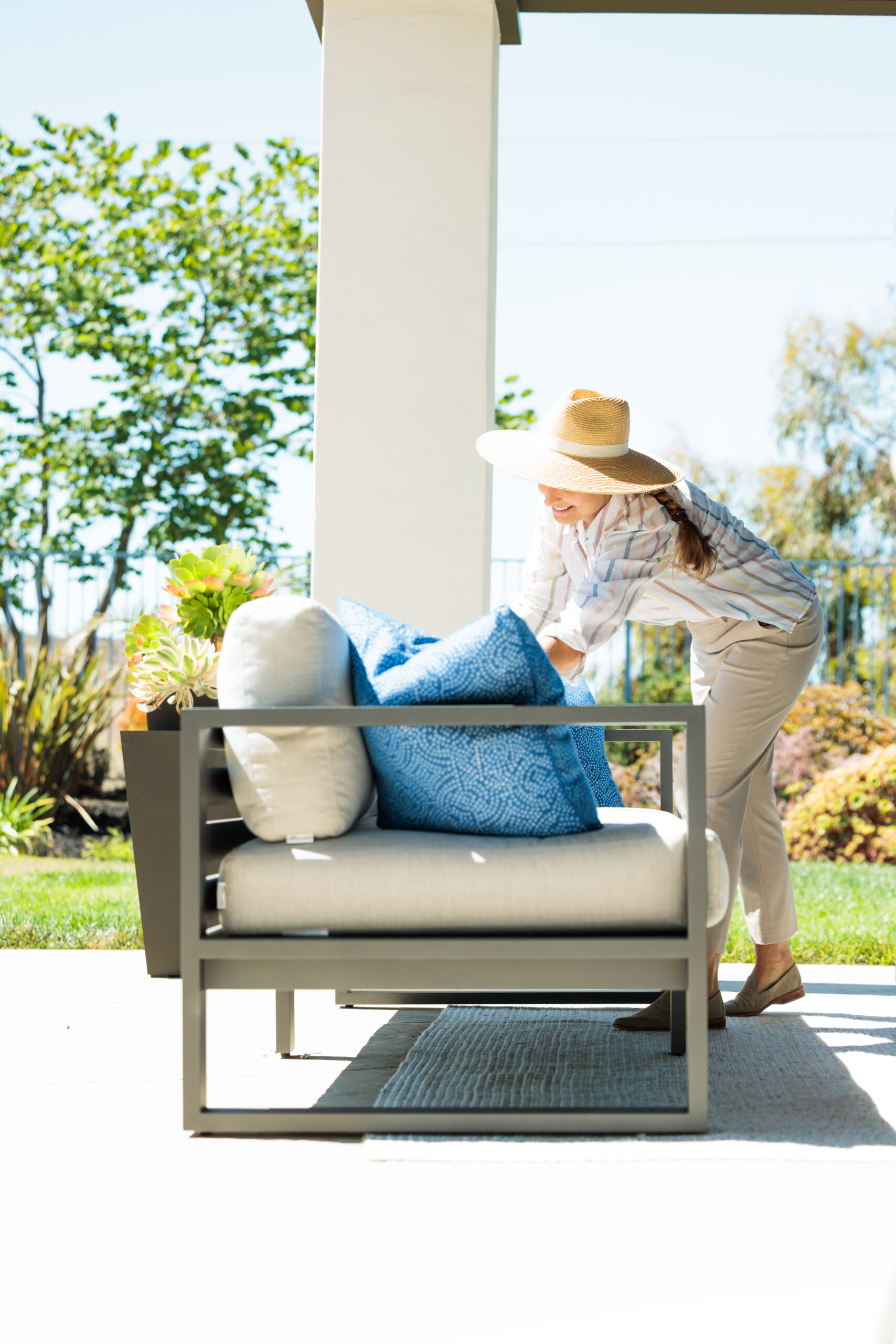 Woman in a Ralph Lauren straw hat fluffing blue and beige pillows on a modern aluminum outdoor couch with green trees in the background.