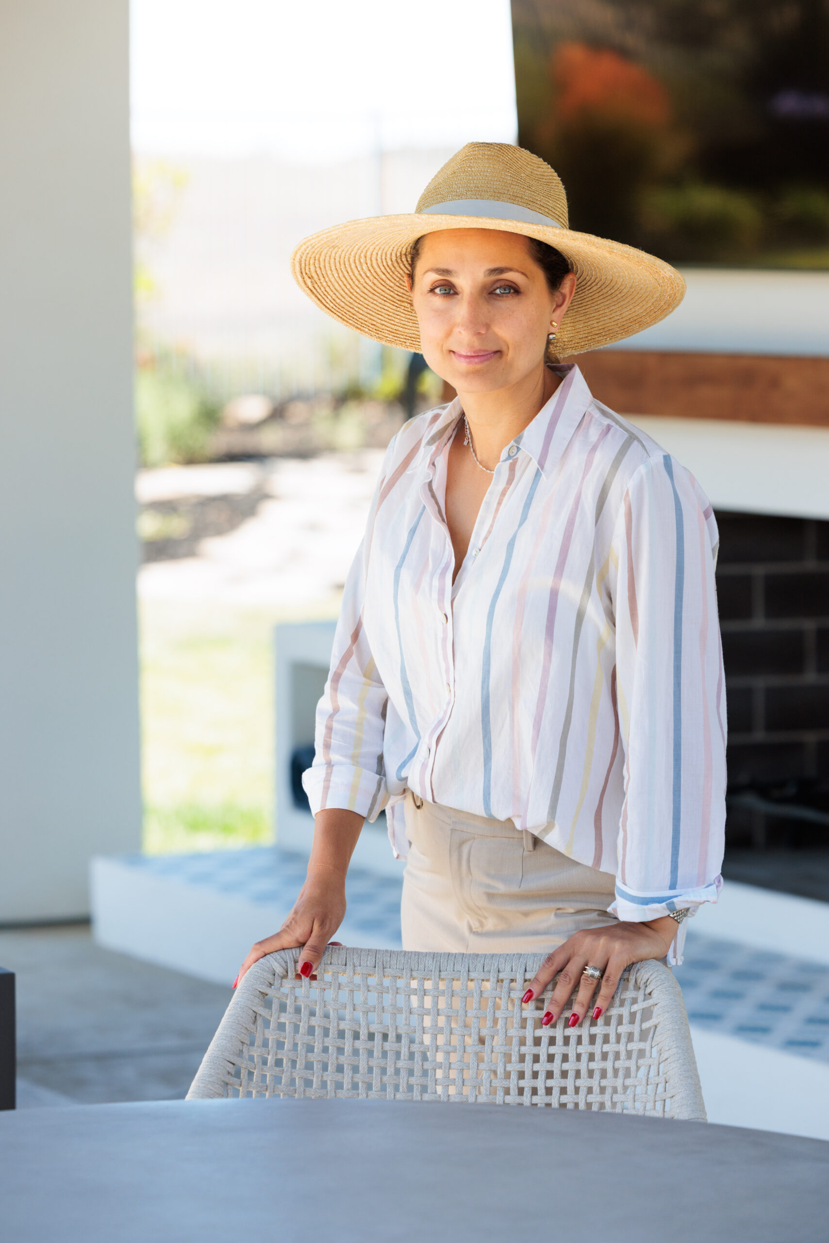 Kristina Jones standing by an outdoor dining table and holding onto a chair, wearing a Ralph Lauren straw hat, light stripped cotton button-down shirt and beige pants. Outdoor fireplace with white stucco in the background with TV hanging on the fireplace and wood mantle under the TV.